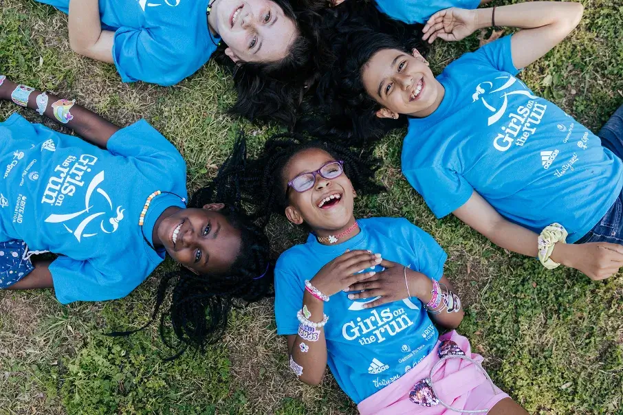 Diverse group of young girls laying in a circle on the ground looking up at the sky smiling and laughing.