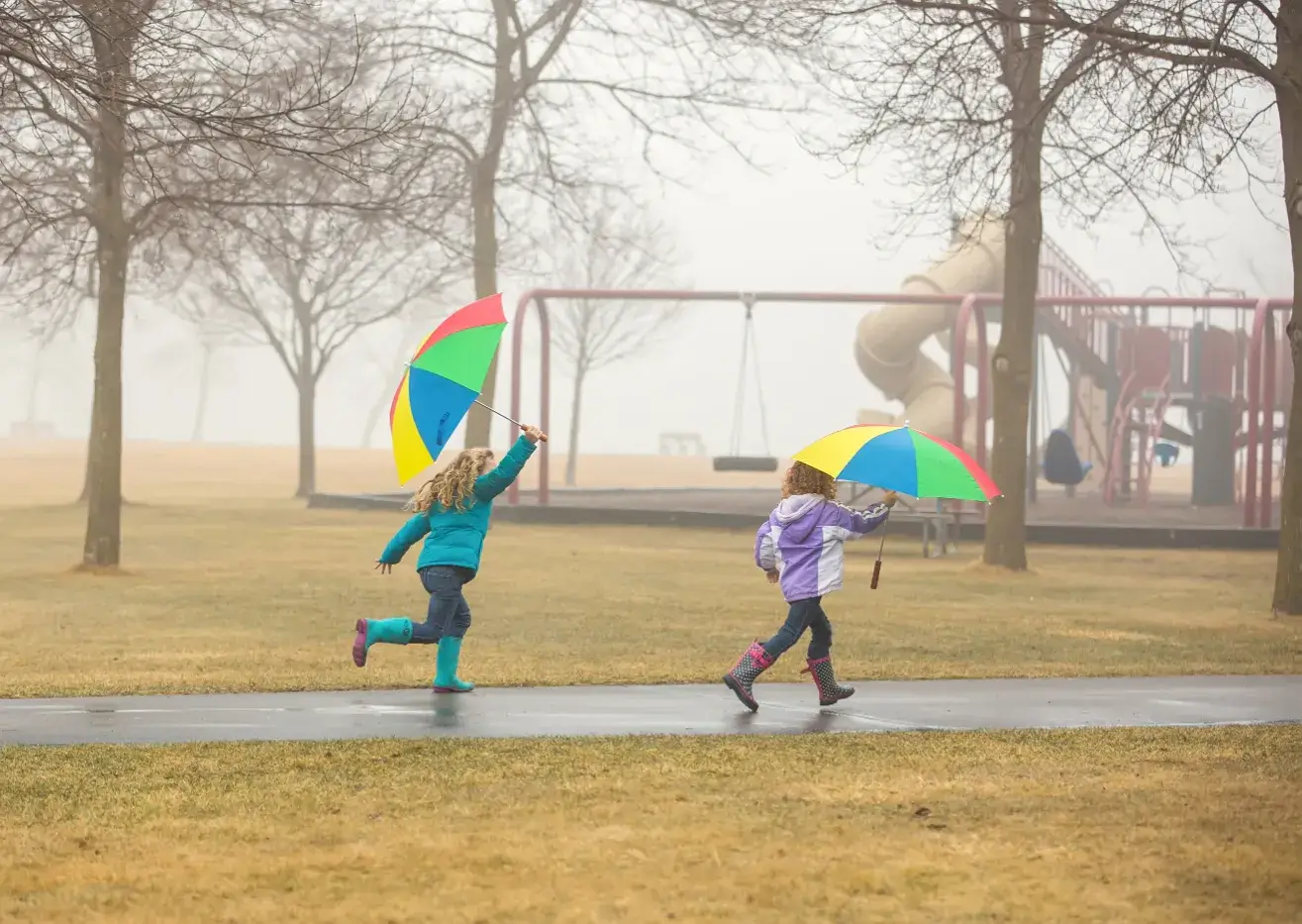 Two young girls at a park running in the rain with umbrellas and rain boots on a foggy day.